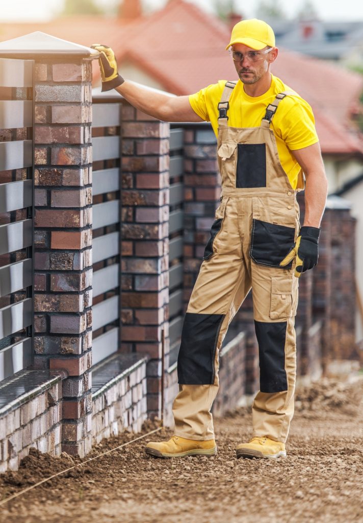 Fence builder in a yellow cap and work overalls with a gloved hand on a brick fence post