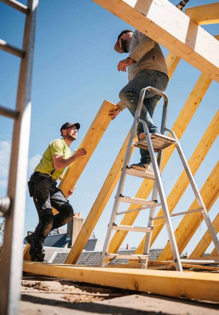 Two framers setting new roof trusses, one on a ladder and one passing up a board