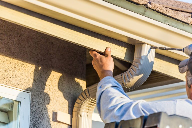 Worker fastening a white gutter downspout under the eaves of a stucco house