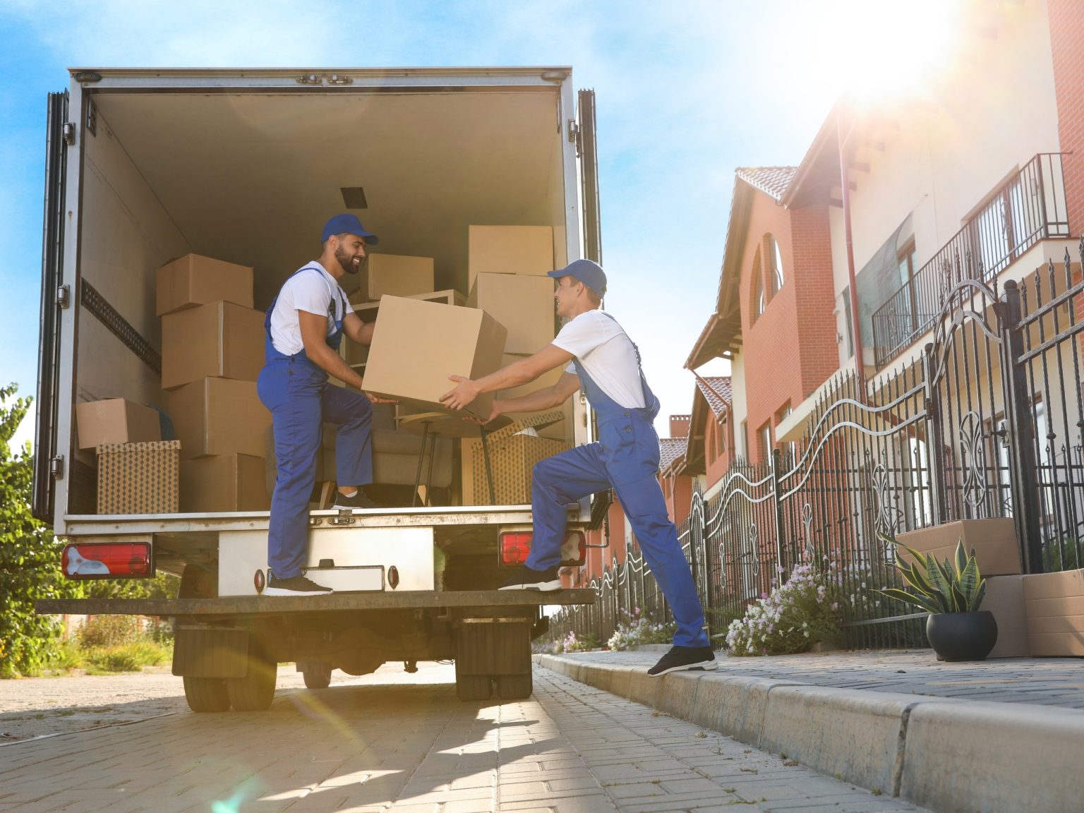 Two movers in blue overalls unloading boxes from the back of a moving truck on a residential street