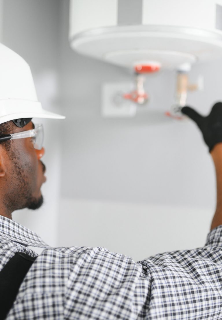 Plumber in a white hard hat and safety glasses reaching up to the valves under a wall-mounted water heater