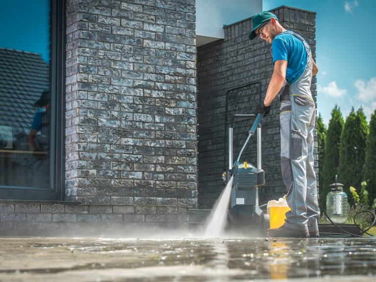 Worker in a green cap pressure washing a stone patio next to a brick wall